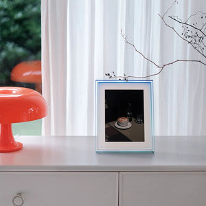 Blue and green ombre frame on a surface with a white curtain and orange chair in the background