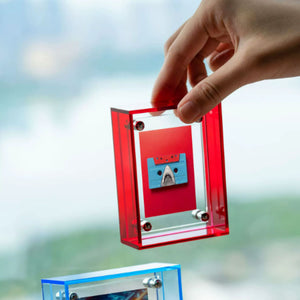 Hand holding a transparent red acrylic picture frame with a blue cassette tape inside against a blurred outdoor background