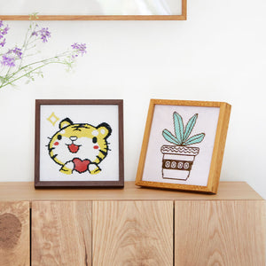 Two framed cross-stitched pieces on a wooden surface with a white wall background. Teak and Black walnut wood square picture frames.