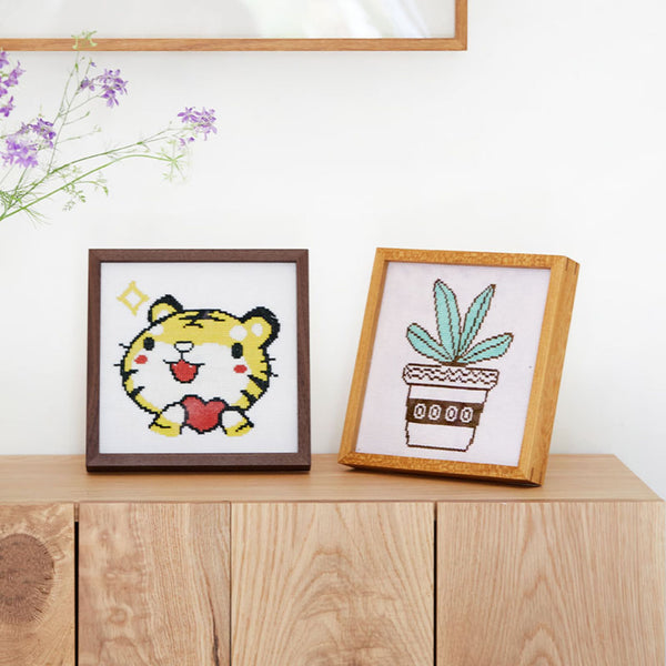 Two framed cross-stitched pieces on a wooden surface with a white wall background. Teak and Black walnut wood square picture frames.