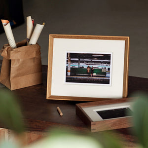 Framed photograph on a wooden surface with a brown paper bag and pencils. Teak and black walnut. 