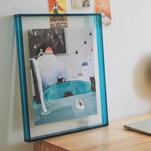 Thick blue picture framed artwork of a snowy scene with a cat and a bird on a desk.