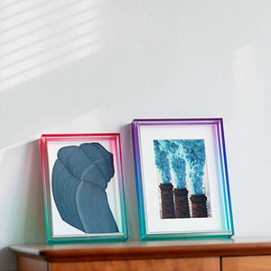 Two ombre acrylic framed artworks on a white wall with a wooden shelf.