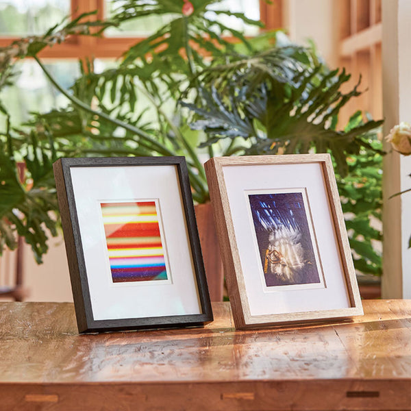 Two framed artworks on a wooden surface with plants in the background. Wenge and white ash wood frame.