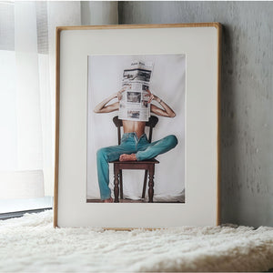 White ash wood framed artwork of a person sitting on a chair reading a newspaper, leaning against a textured wall.