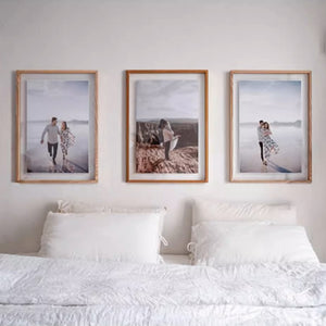 Three framed photographs of a couple on a beach above a bed with white bedding. Two white ash wood plus one teak wood.