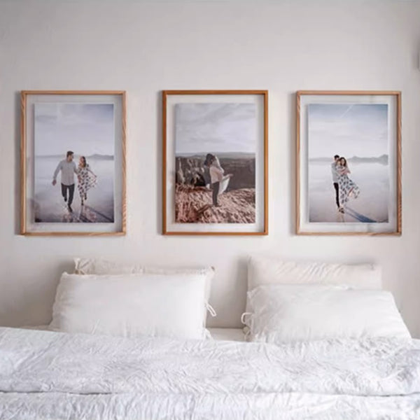 Three framed photographs of a couple on a beach above a bed with white bedding. Two white ash wood plus one teak wood.