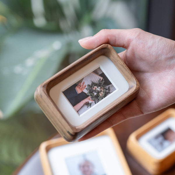 Person with hand holding a small black walnut wooden picture frame with a photo of a baby on a blurred green background