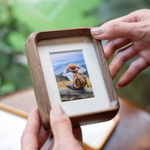 Person holding a black walnut photo frame with a scenic picture of a person in a hat and backpack.
