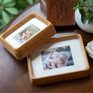 Wooden photo frame with photos of a dog and two people on a wooden surface with plants in the background.