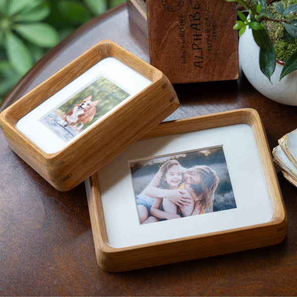 Wooden photo frame with photos of a dog and two people on a wooden surface with plants in the background.